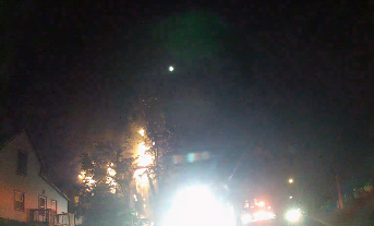 Dark night in a neighborhood. Emergency light glares. Fire visible through trees.